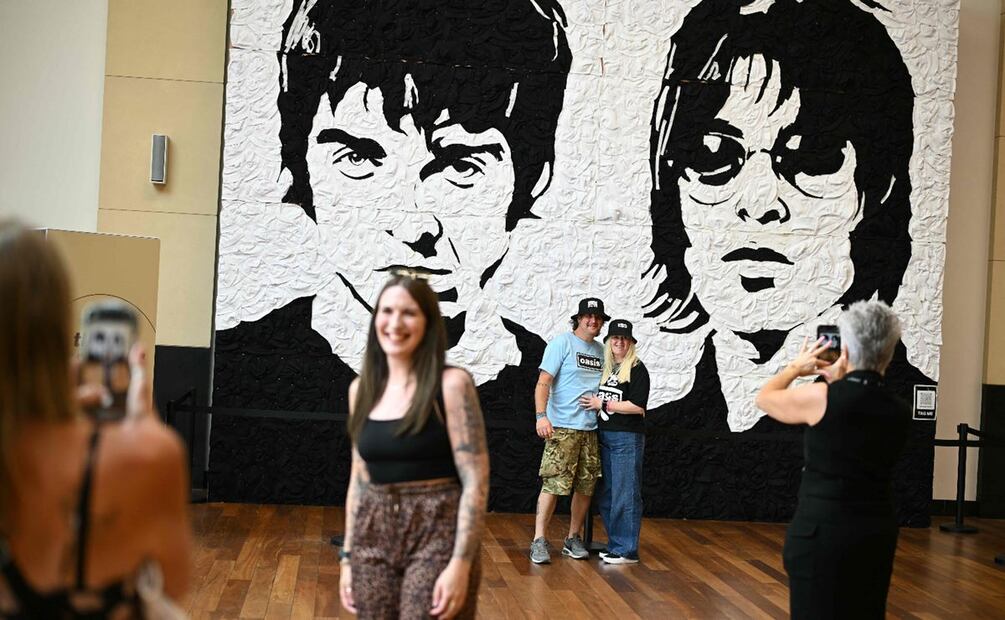La gente posa para una foto frente a una enorme obra de arte de la banda de rock británica Oasis, llamada "The Wonder Wall", creada por Nathan Wyburn con sombreros de pescador en Cardiff el 4 de julio de 2025, antes del concierto inaugural de su esperadísima gira de reunión, casi 16 años después de su última actuación juntos. 
Foto: Oli SCARFF / AFP.